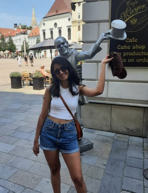 A girl posing for a photo while imitating a statue of a man greeting with his cap.