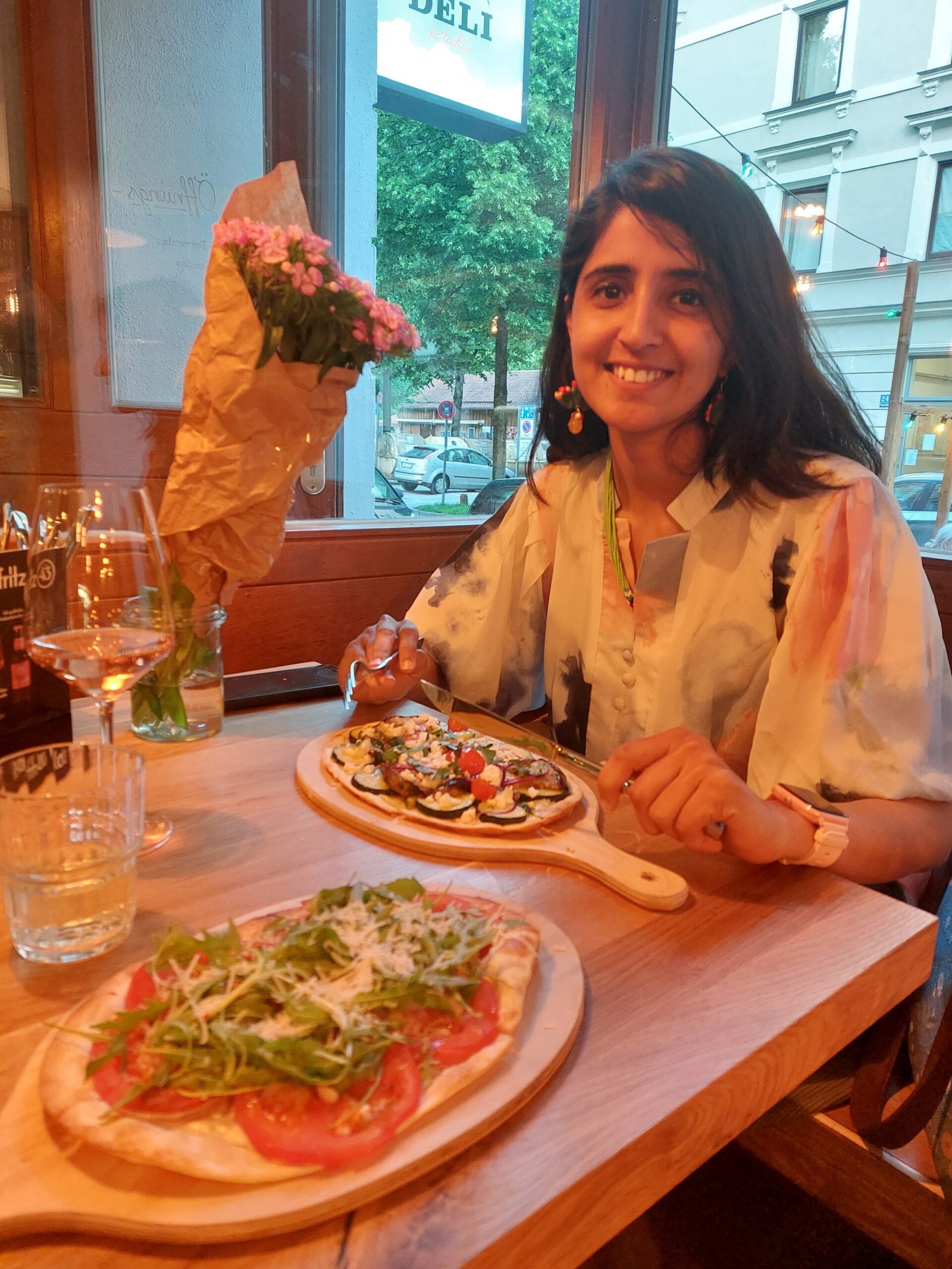 A smiling woman sits by a café window with a bouquet beside her, enjoying colorful flatbreads and drinks in a warm, relaxed setting.
