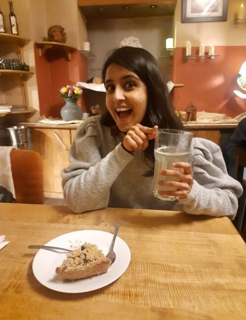 A smiling woman points toward a dessert while holding a glass at a restaurant table; warm lighting and colorful décor create a cheerful, relaxed mood.