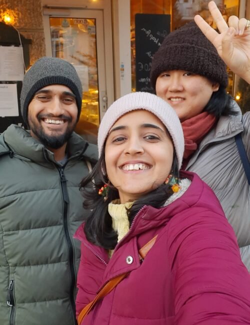 Three smiling friends in winter coats and knit hats pose for a selfie outside a warmly lit café; one raises a peace sign, creating a cheerful, cozy mood.