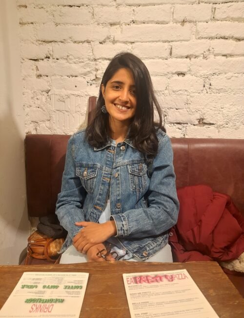 A smiling woman in a denim jacket sits at a wooden café table with menus, against a white brick wall; a red coat beside her adds a relaxed, welcoming feel.