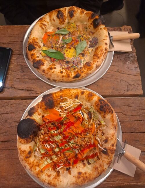 Two wood-fired pizzas on metal plates atop a rustic wooden table: one topped with leafy greens and cheese, the other with vegetables and shredded toppings.