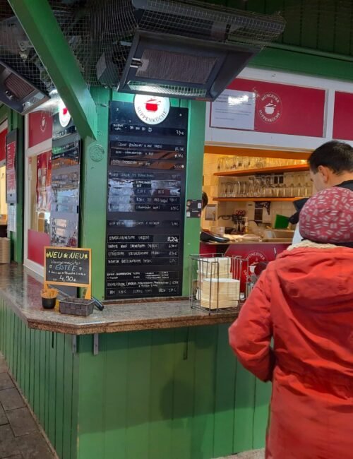 A customer in a red jacket and knit cap orders at a green food stall beneath menu boards, amid a busy, warmly lit indoor market.