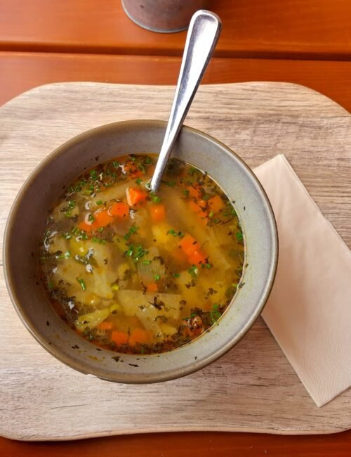 A warm bowl of vegetable soup with carrots, herbs, and diced vegetables sits on a wooden tray beside a folded napkin, evoking a cozy meal.