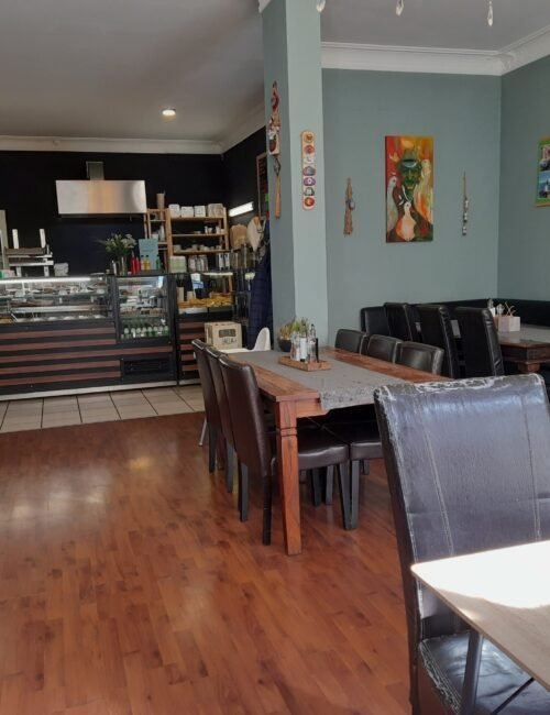 A quiet café interior with a wood-floored dining area, dark tables and chairs, and a glass-fronted counter stocked with food and drinks.