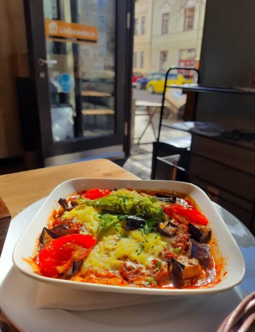 A square white bowl of baked vegetables topped with melted cheese and herbs sits on a café table, with red peppers and eggplant in a cozy street-side setting.