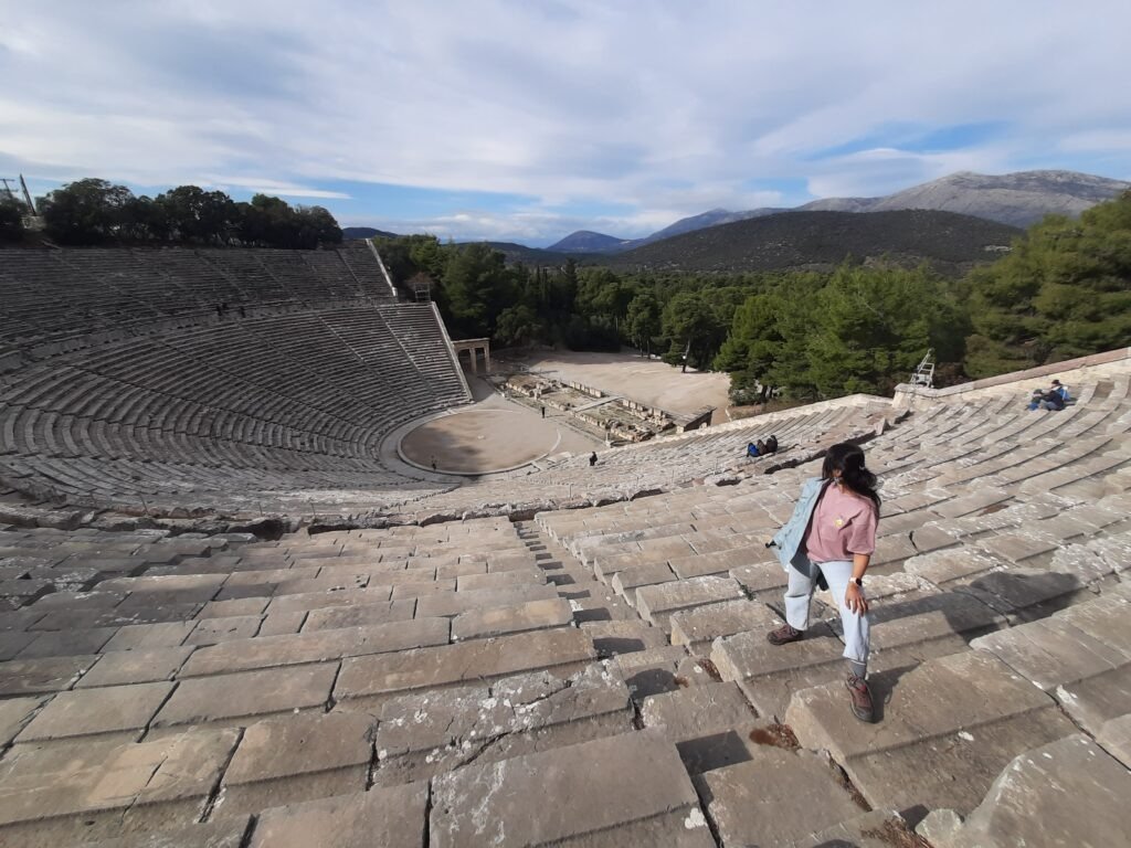 A woman walks down the weathered stone seats of an ancient open-air amphitheater, with green forest, distant mountains, and a cloudy blue sky beyond.
