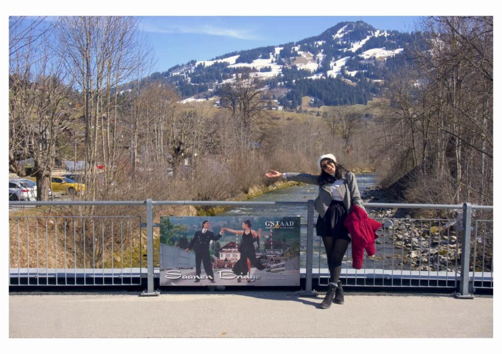 A woman in a knit hat points toward a river and snowy mountain from a bridge, with leafless trees and a scenic sign nearby; a calm winter outing.