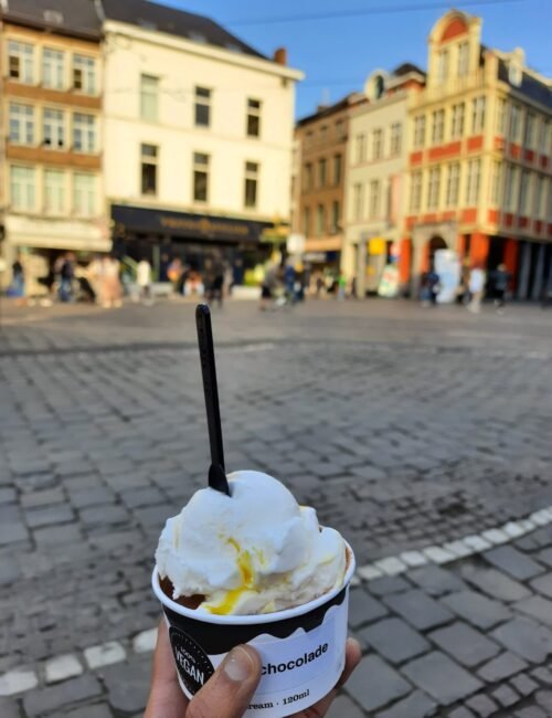 Hand holds a cup of white and yellow ice cream with a spoon in a sunny cobblestone square, framed by colorful historic buildings.