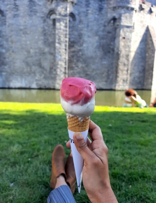 A hand holds a pink-and-white ice cream cone before a weathered stone castle, with green grass and a seated person in the sunny background.