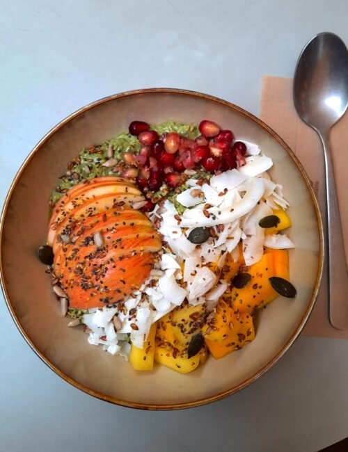 Overhead view of a vibrant smoothie bowl topped with mango, papaya, kiwi, pomegranate, coconut, seeds, and carrot, beside a spoon on a table.