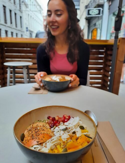 Smiling woman at a café table holds a grain bowl while a colorful bowl with apples, rice, pomegranate, and vegetables fills the foreground.
