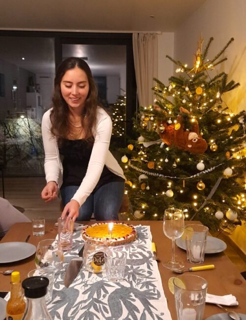 Young woman lights a candlelit dessert at a dining table beside a decorated Christmas tree, creating a warm, festive holiday scene.