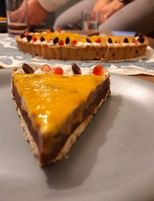 Close-up of a glossy yellow fruit tart slice on a plate, with a berry-topped tart and diners in the background, creating a cozy mood.