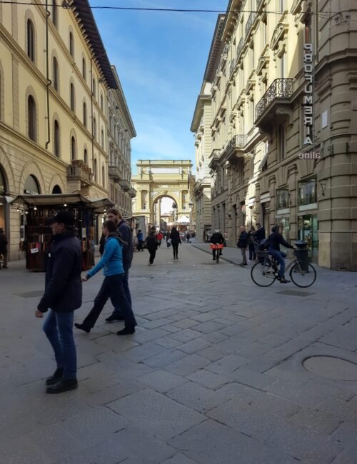Historic Florence street with pedestrians and cyclists, elegant arcaded buildings and shops, and a sunlit stone arch framing the view.