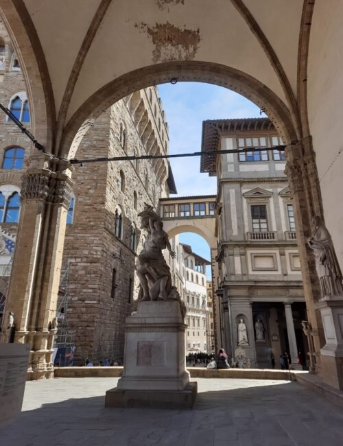 Historic Italian plaza framed by stone arches, featuring a central marble statue, medieval buildings, bright blue sky, and pedestrians in sunlight.