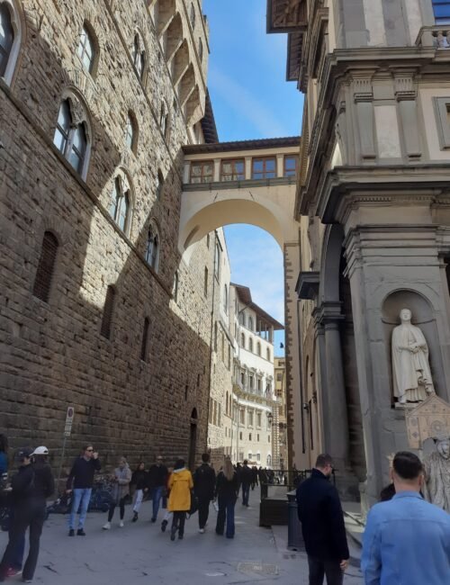 Pedestrians stroll through a historic Italian street beneath an arched skybridge, flanked by weathered stone walls and an ornate church façade.