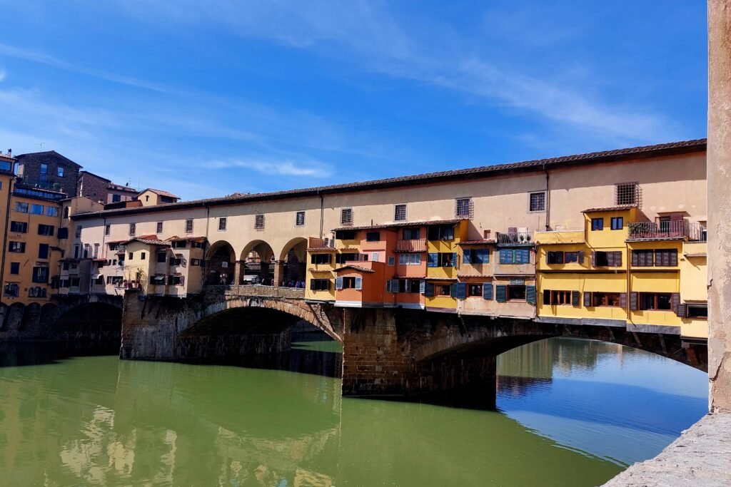 Historic Ponte Vecchio in Florence spans the green Arno River, lined with colorful shops and arched stone supports beneath a blue sky.