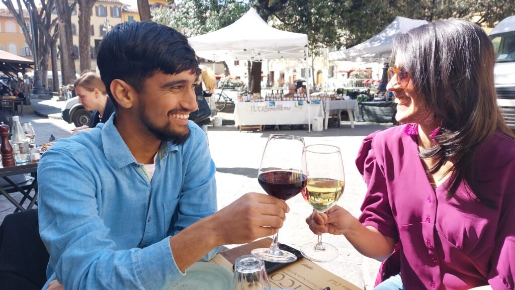 Smiling couple clinks red and white wine glasses at an outdoor market café, with sunny stalls and trees creating a relaxed, festive mood.
