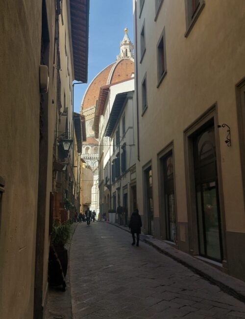 Narrow historic alley in Italy, lined with ochre buildings and cobblestones, leading toward a domed church tower under a blue sky.