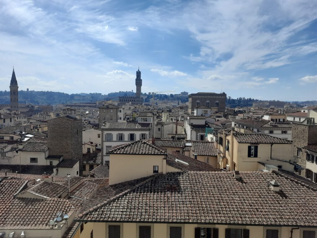 Panoramic Florence cityscape with terracotta rooftops, historic towers and domes beneath a bright blue, cloud-streaked sky.