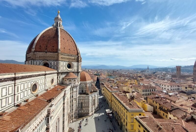 Florence skyline with Brunelleschi’s red-tiled dome and cathedral in foreground, viewed above sunlit streets and historic rooftops under blue sky.