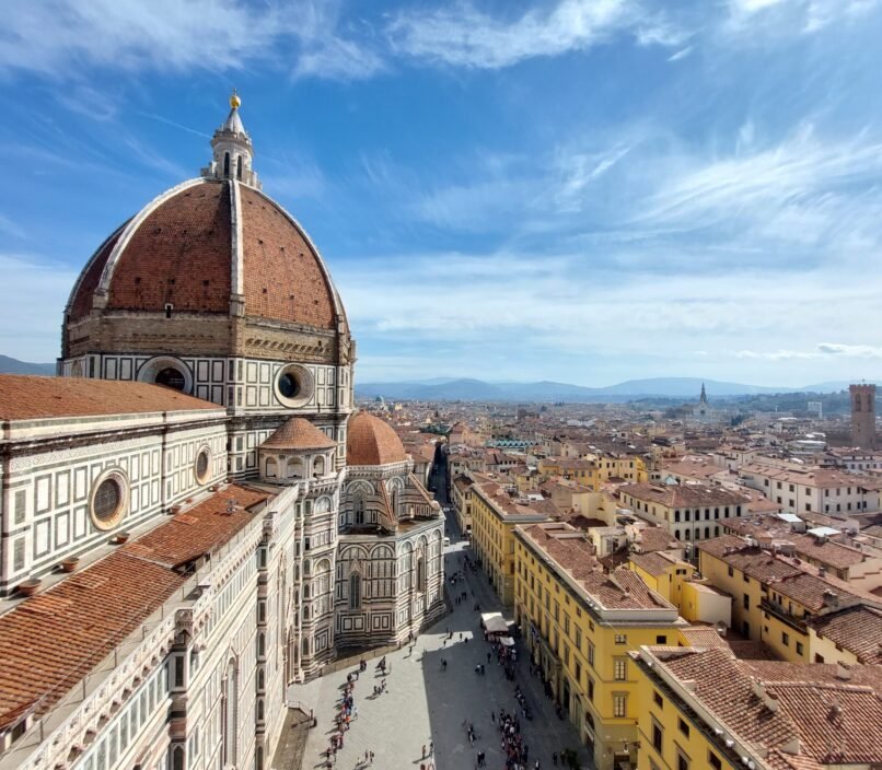Florence skyline with Brunelleschi’s red-tiled dome and cathedral in foreground, viewed above sunlit streets and historic rooftops under blue sky.