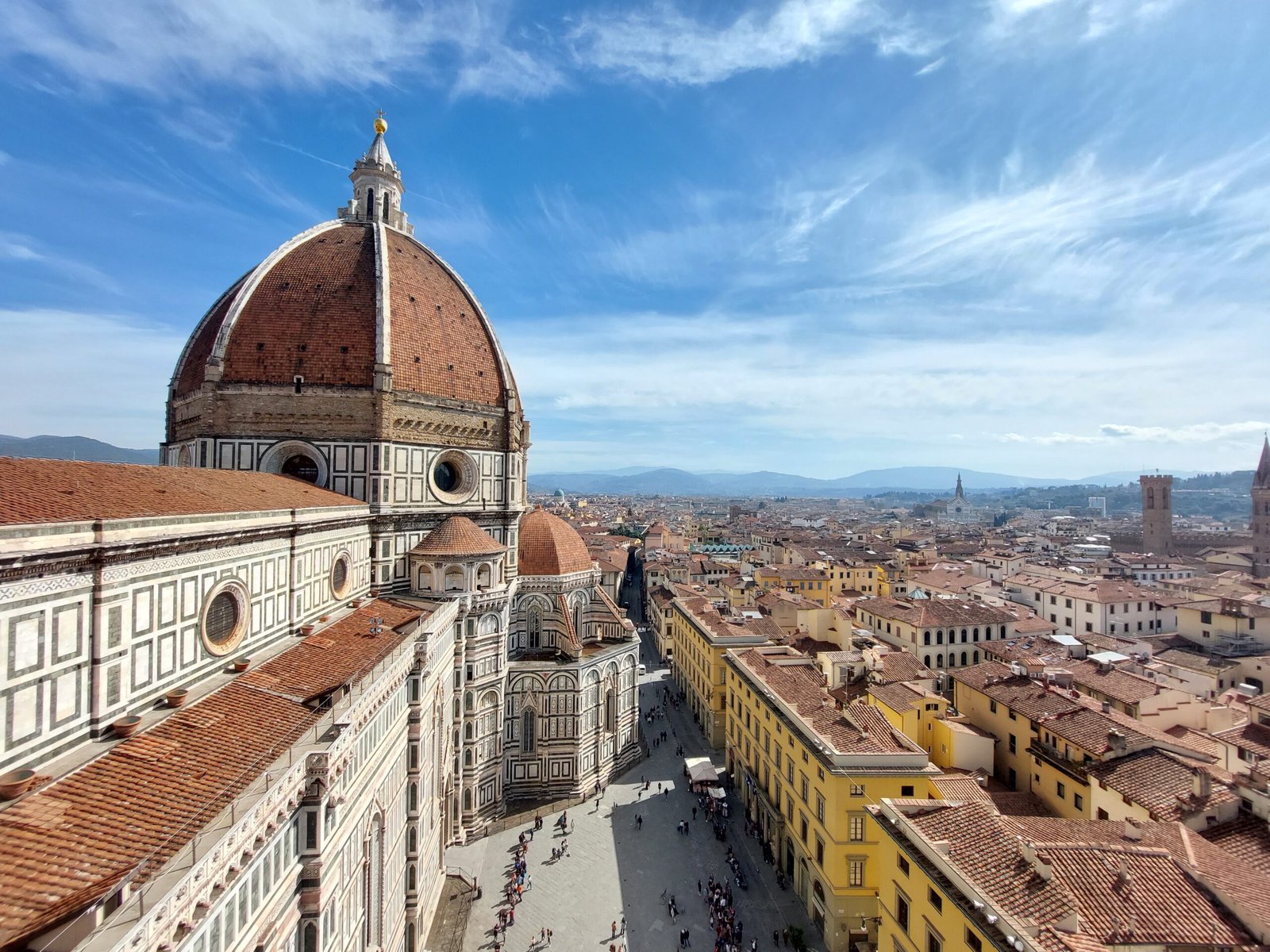 Florence skyline with Brunelleschi’s red-tiled dome and cathedral in foreground, viewed above sunlit streets and historic rooftops under blue sky.
