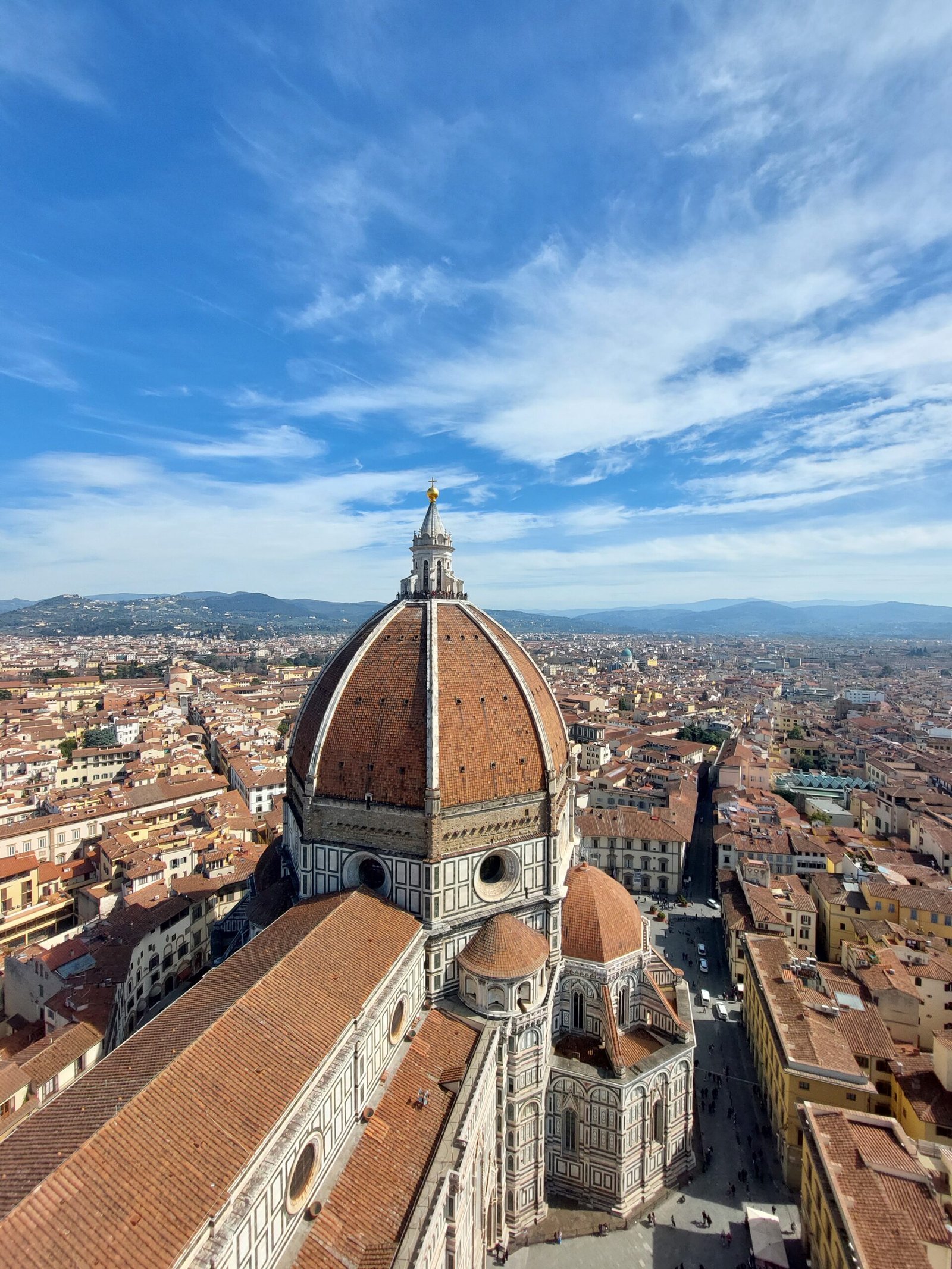 Florence’s Duomo with its iconic red-tiled dome overlooks terracotta rooftops and streets under a bright blue sky, creating a historic cityscape.