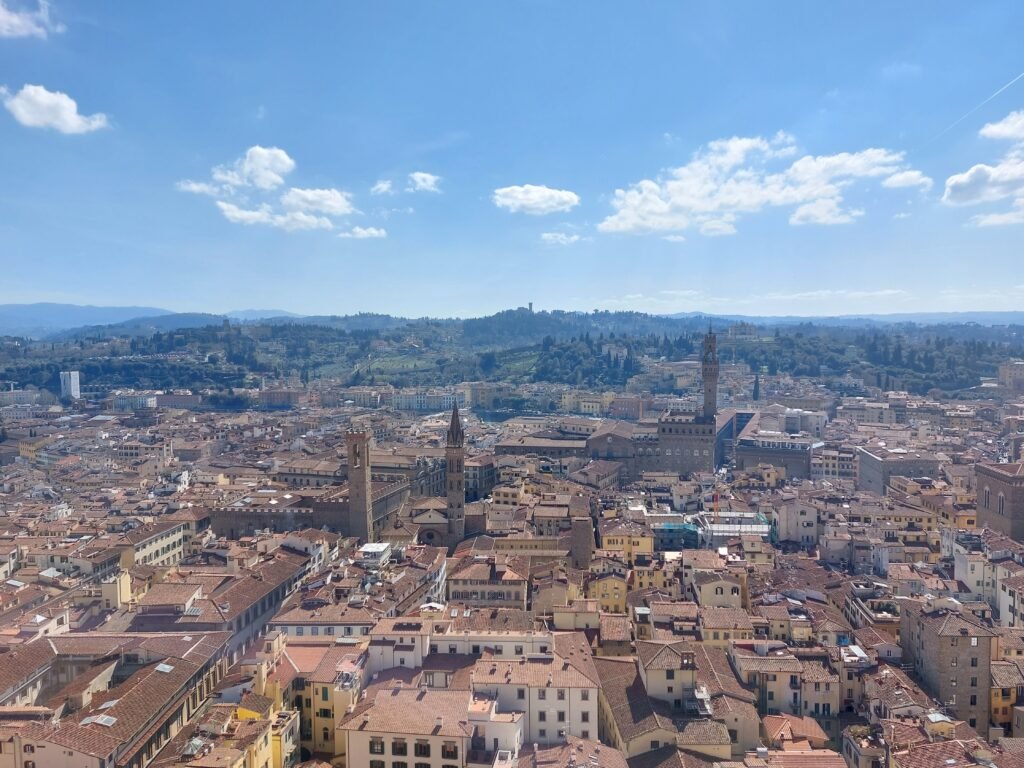 Panoramic view of Siena, Italy, with dense terracotta rooftops, historic towers, rolling hills, and a bright blue sky creating a serene scene.