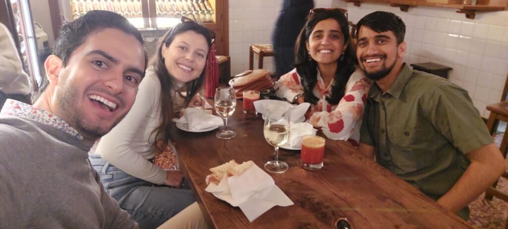 Four friends smile for a selfie at a cozy restaurant table, surrounded by plates, napkins, wine glasses, and a candle, conveying warmth and joy.
