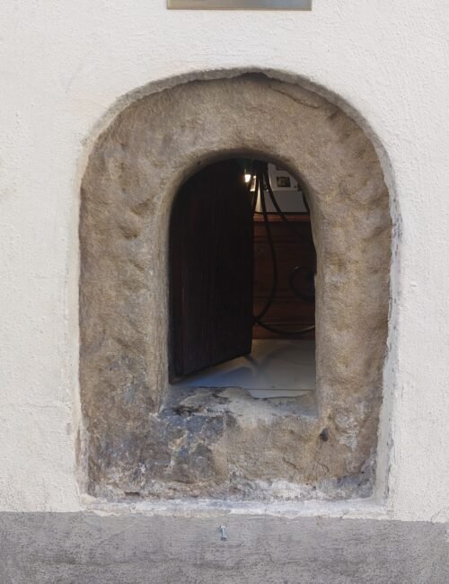 Rustic stone-arched wine window set in a white plaster wall, with a “Buchetta di Vino” sign and dark wooden opening creating historic charm.