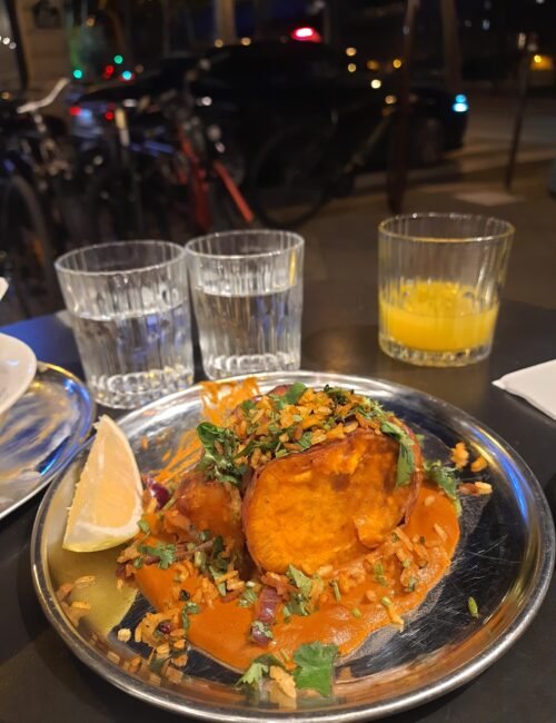 Restaurant table with a plated golden-brown dish in orange sauce, herbs and lemon wedges, water glasses, and orange juice; cozy night setting.