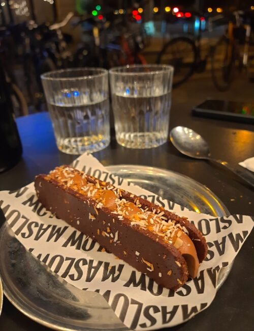 Chocolate-covered pastry topped with shredded coconut on a café plate, with water glasses, spoon, bicycles, and colorful night lights behind it.