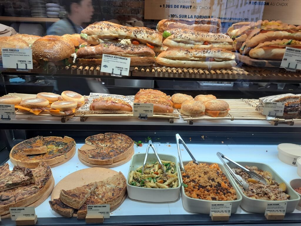 Bakery counter displaying stacked sandwiches, crusty breads, pastries, quiches, and prepared salads in glass cases; an abundant, inviting spread.