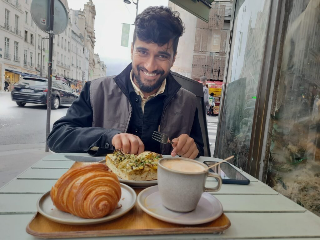 Smiling man at a Parisian café table with a croissant, savory toast, and cappuccino, creating a warm, inviting breakfast scene.