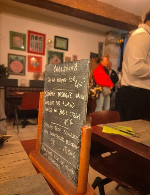 Handwritten chalkboard menu in a cozy café, listing desserts and drinks; colorful wall art, tables, and casually dining people create a warm mood.