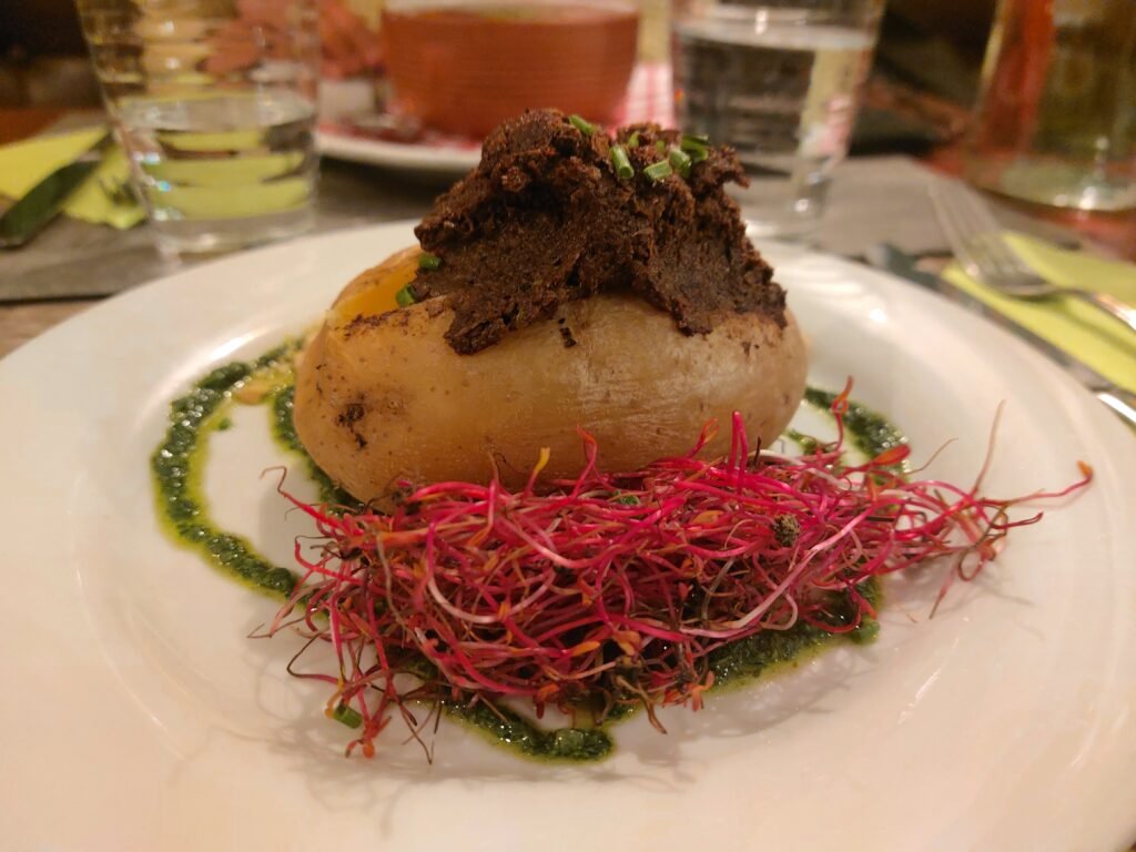 Gourmet roasted potato topped with dark crumb and herbs, surrounded by bright pink beet sprouts and green sauce on a white plate.