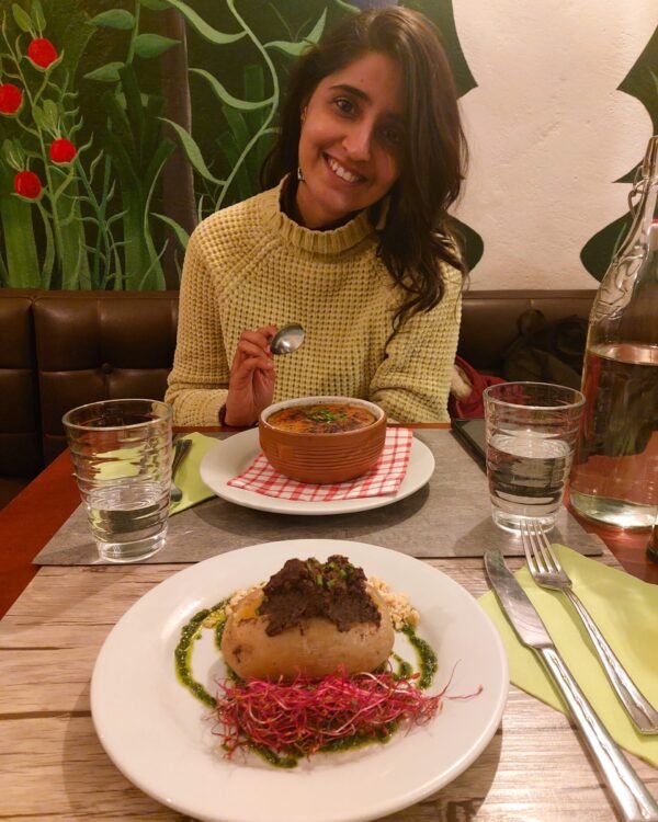 Smiling woman holds a spoon beside a bowl of soup and plated gourmet dish, seated at a restaurant table with drinks and floral mural.