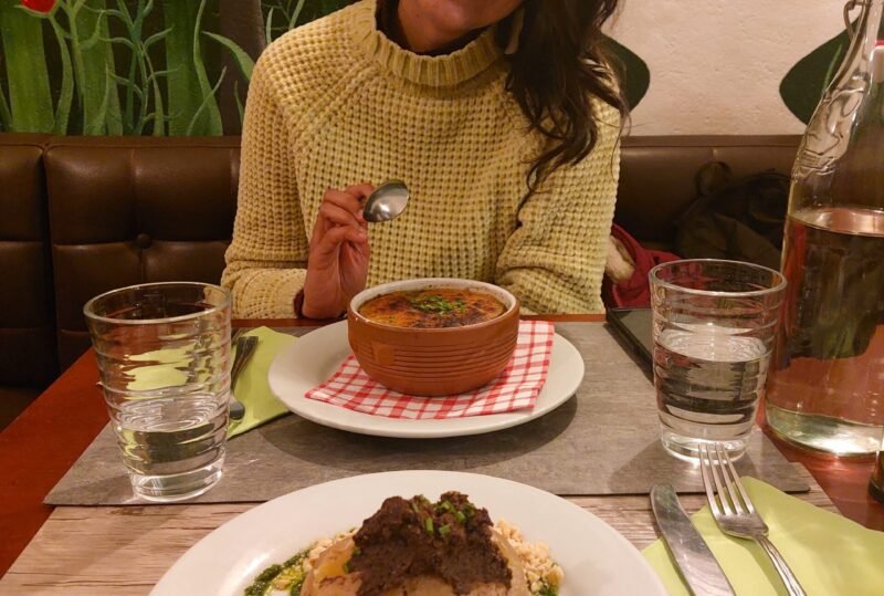Smiling woman holds a spoon beside a bowl of soup and plated gourmet dish, seated at a restaurant table with drinks and floral mural.