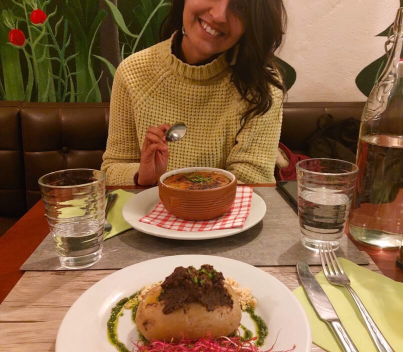 Smiling woman holds a spoon beside a bowl of soup and plated gourmet dish, seated at a restaurant table with drinks and floral mural.
