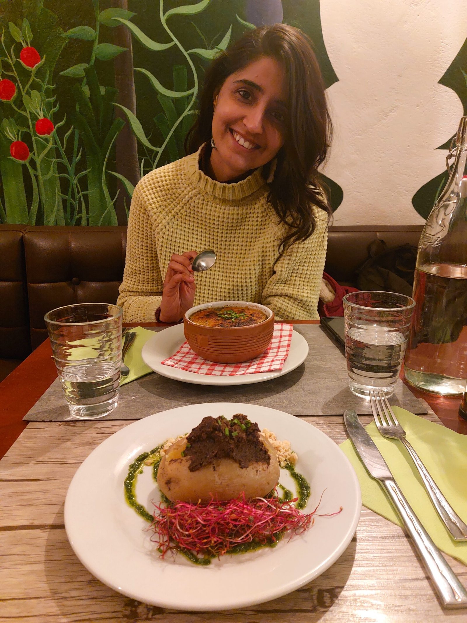 Smiling woman holds a spoon beside a bowl of soup and plated gourmet dish, seated at a restaurant table with drinks and floral mural.