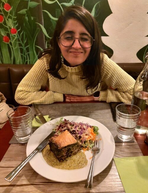 Smiling woman in a cream sweater and glasses sits at a cozy restaurant table behind salmon, vegetables, and sauce on a white plate.