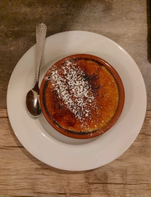 Top-down view of a caramelized crème brûlée dusted with powdered sugar in a brown ramekin on a white plate, with a spoon beside it.