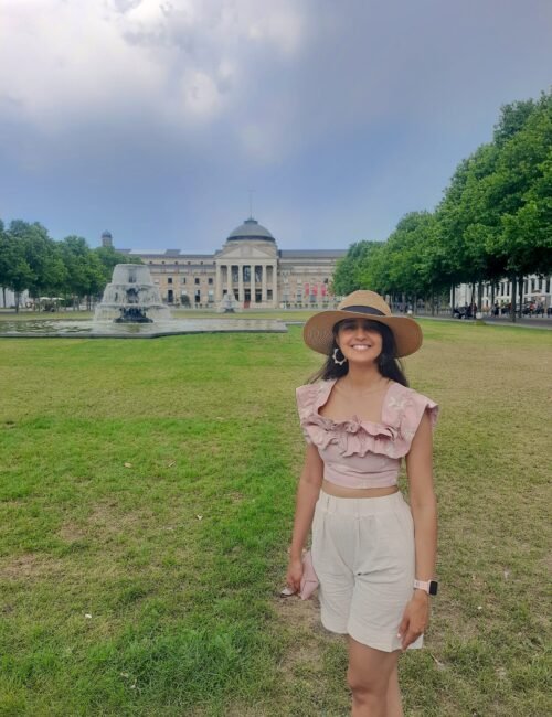 A woman dressed in light pink top and beige shorts posing in front of a historic building and a water fountain.