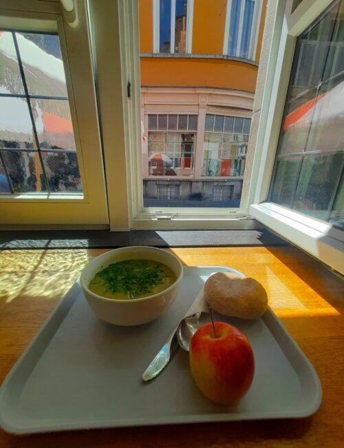 Sunlit breakfast tray by an open window holds herb-garnished soup, bread, red apple, and spoon; colorful city buildings create a warm mood.