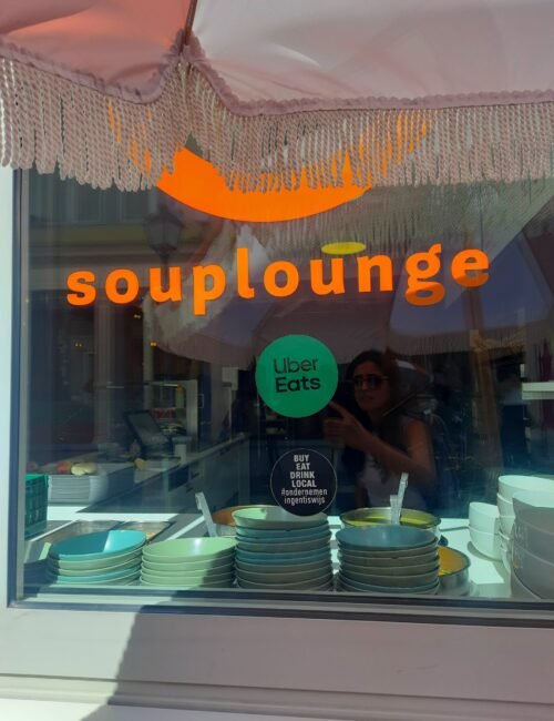 Restaurant window displaying the orange “soup lounge” sign, Uber Eats sticker, colorful stacked dishes, and scalloped awning.