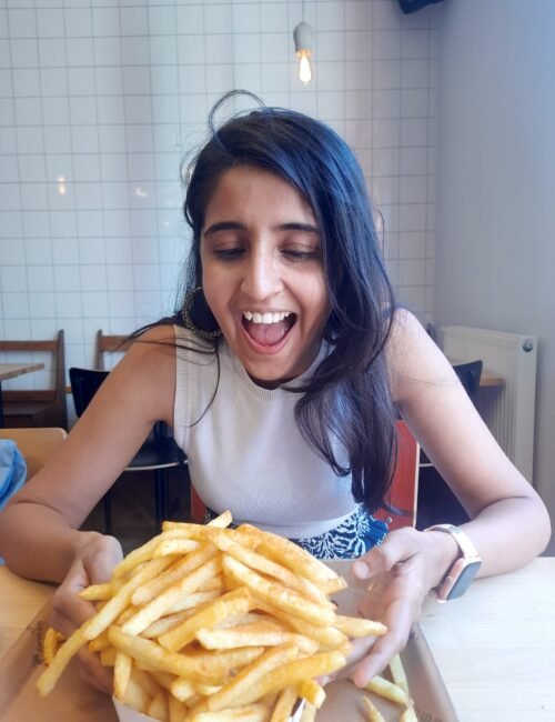 Joyful woman with long dark hair laughs at a huge pile of golden French fries on a table, conveying surprise and delight in a cozy café.