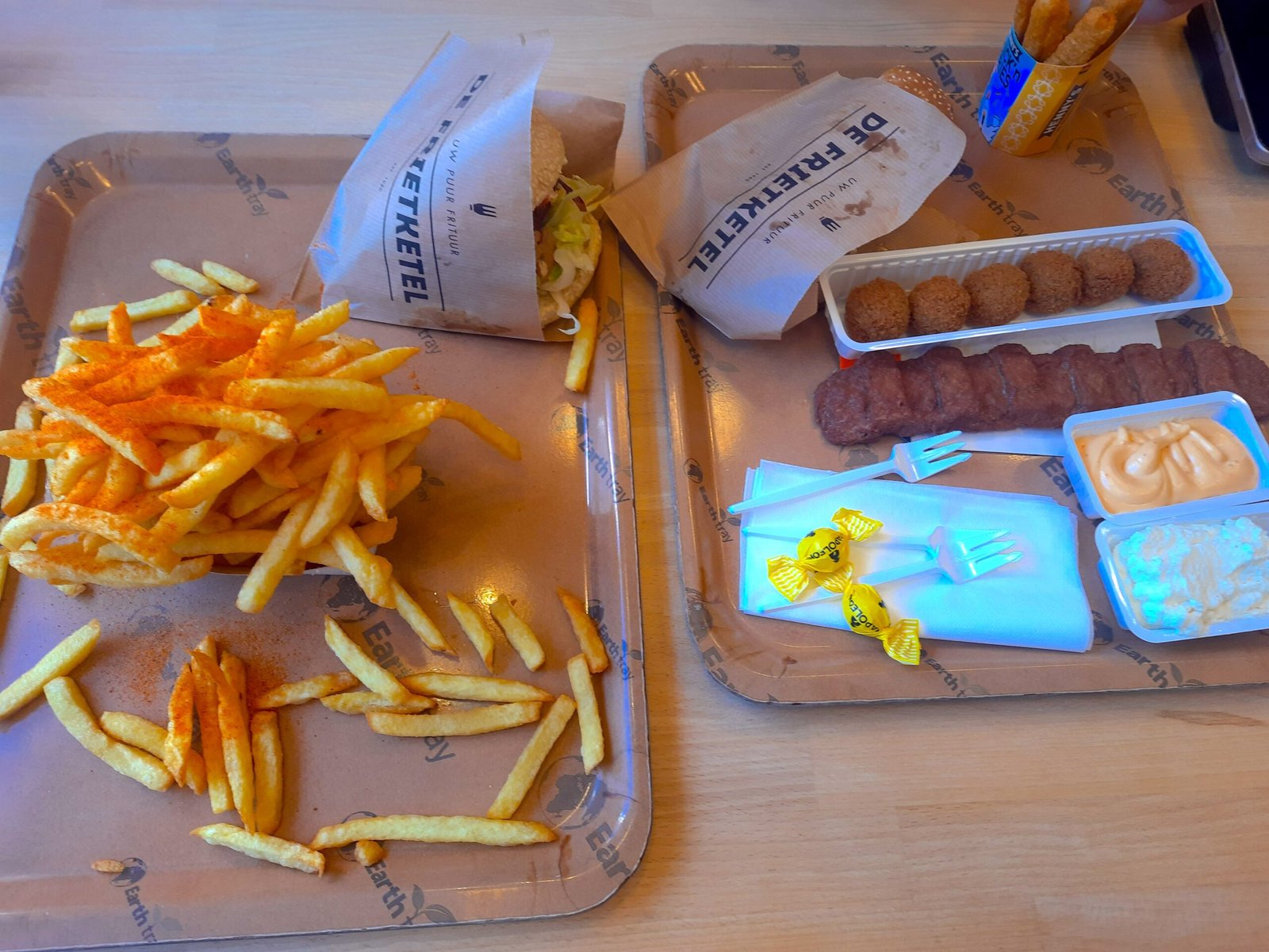 Fast-food spread on trays: overflowing fries, burgers in paper wrappers, croquettes, sausages, dipping sauces, forks, and wrapped candies.