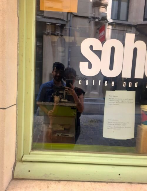Lime-green café window reflecting two people taking a photo, with oversized white “son” lettering, “Coffee and” text, and a menu.